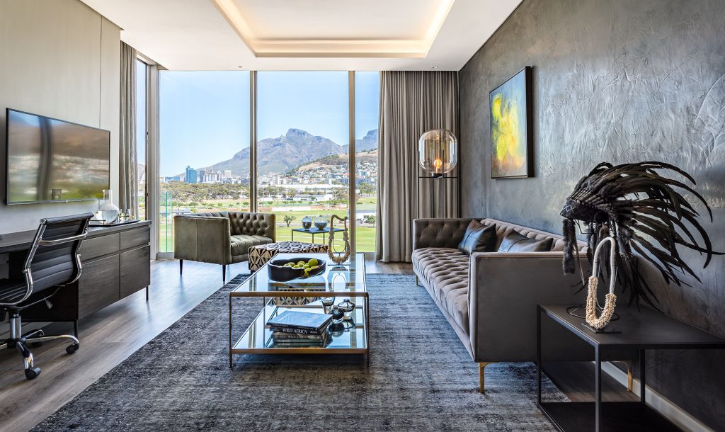 Ein luxuriöses Wohnzimmer mit dunklen Oberflächen und bodentiefen Fenstern, die auf einen Golfplatz hinausblicken. Der Raum verfügt über eine Couch, einen Couchtisch, eine Lampe, einen Fernseher, einen Arbeitsplatz und eine Leinwand an der Wand.